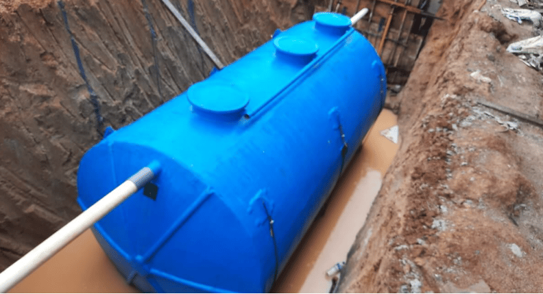 Blue septic tank being installed in an excavation, with access lids and a pipe visible.