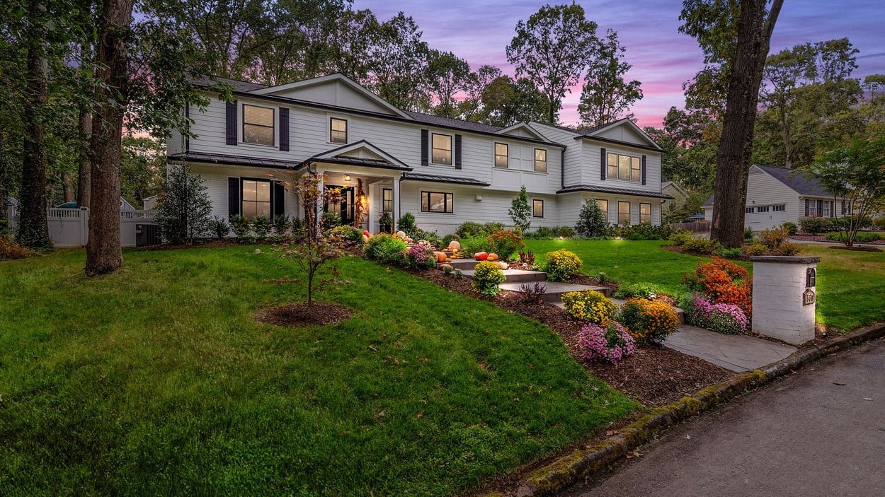 White two-story house with dark shutters, pumpkin-decorated entry, and colorful front garden at dusk.