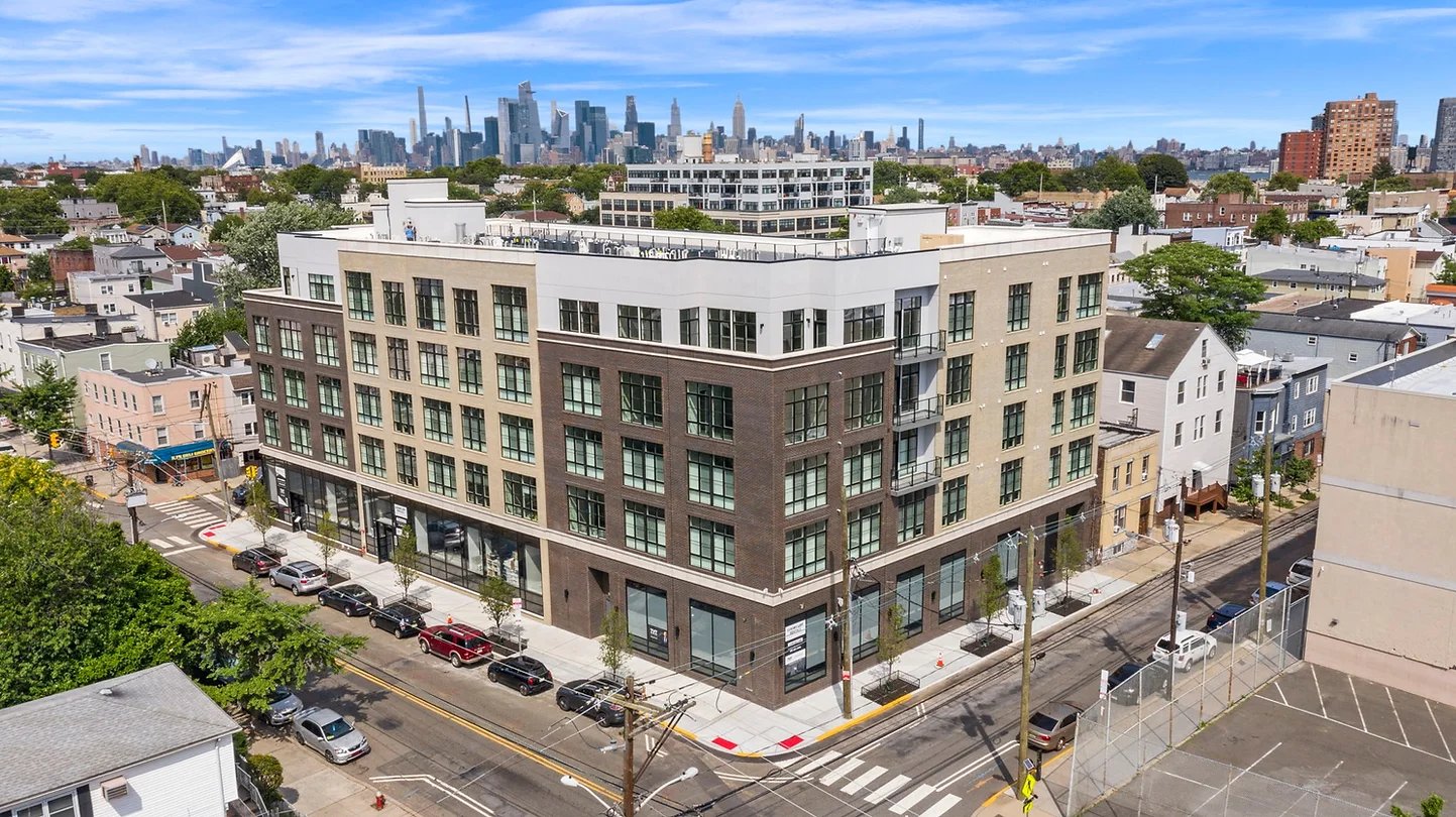 Modern corner apartment building with brick façades and a city skyline in the distance.