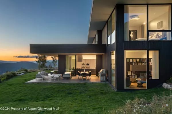 Modern glass-walled home at dusk with a cantilevered roof, furnished patio, and distant mountain views.