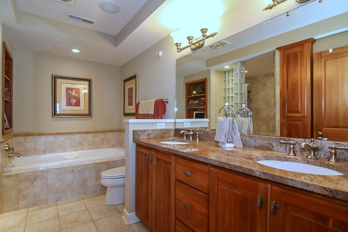 Penthouse bathroom with double granite vanity, wood cabinetry, and a tiled bathtub.