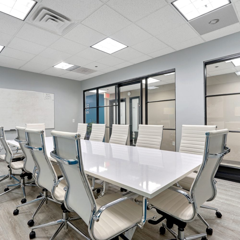 Office conference room with a glossy white table, padded chairs, glass partitions, and a wall-mounted whiteboard.