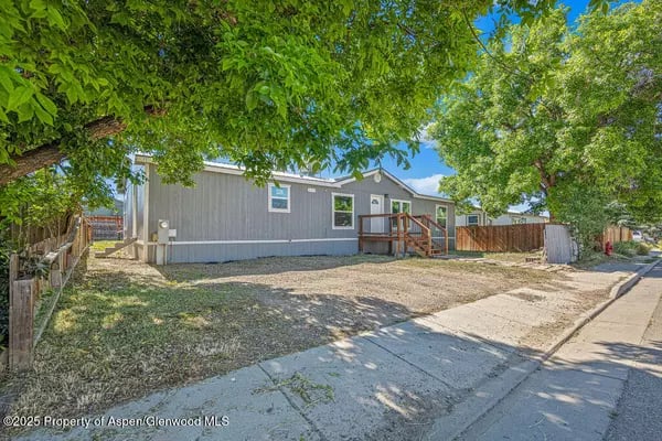 Gray single-story home shaded by leafy trees, with a raised wooden entry deck beside the sidewalk.