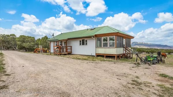 Rural home with a green metal roof and raised porch beside a dirt driveway, with mountains in the distance.