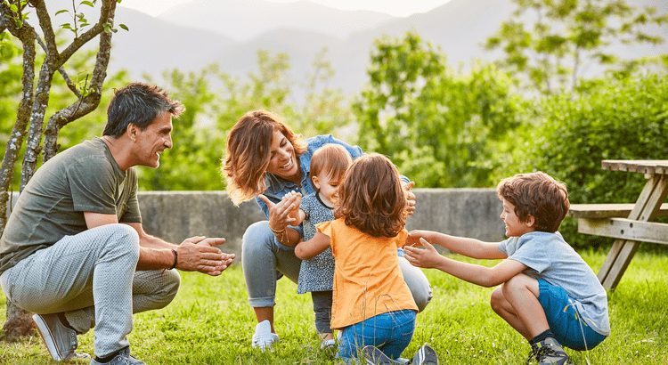A joyful family of five plays on a sunny lawn. Parents kneel, smiling as three children run toward them. Greenery and a picnic table are in the background.