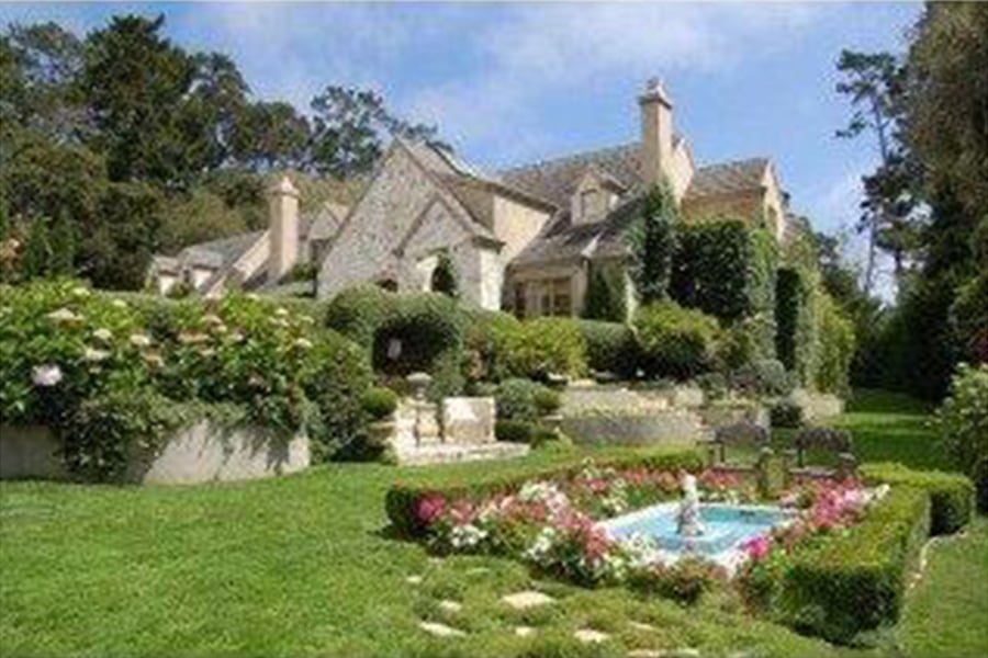 Stone house framed by landscaped gardens, with a flower-bordered fountain and broad lawn.