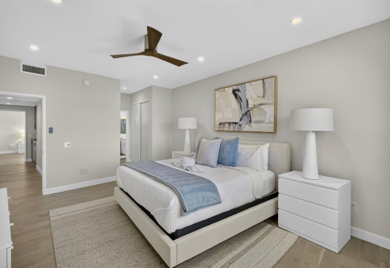 Staged bedroom with a white bed, blue bedding, abstract wall art, and a dark wood ceiling fan.