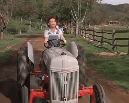 A woman drives a vintage tractor down a dirt path, surrounded by trees and a rustic fence in a scenic countryside setting.