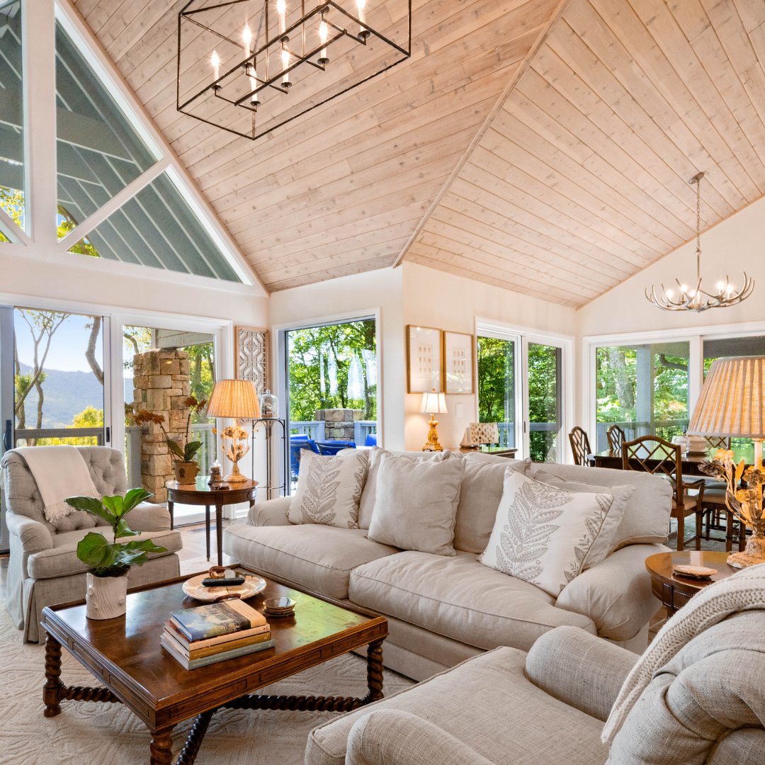 Living room with a vaulted wood ceiling, triangular windows, and views of trees and distant hills.