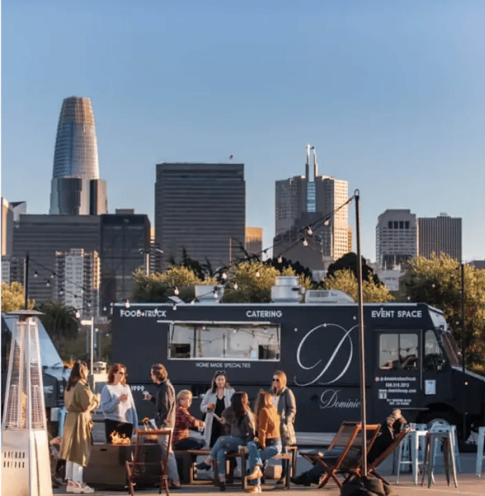 Group of people socializing at outdoor seating area with dark food truck and patio heater, San Francisco skyline including Salesforce Tower visible across water in golden hour light