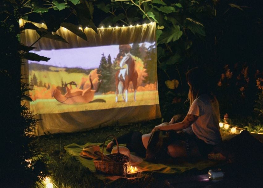 A child watches an animated movie projected outdoors, surrounded by string lights and picnic blankets.
