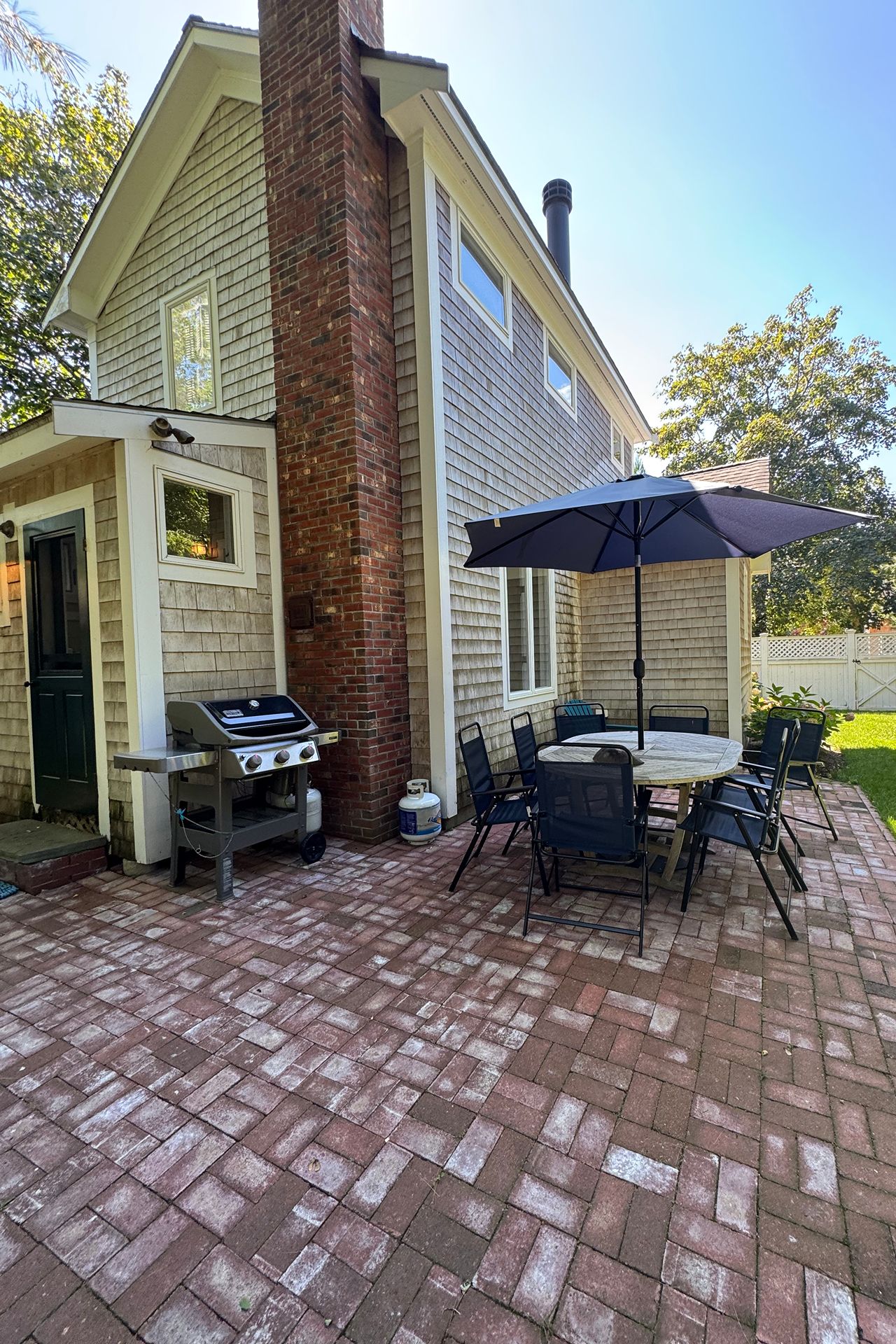 Shingled home beside a brick patio with outdoor dining set, umbrella, and grill.
