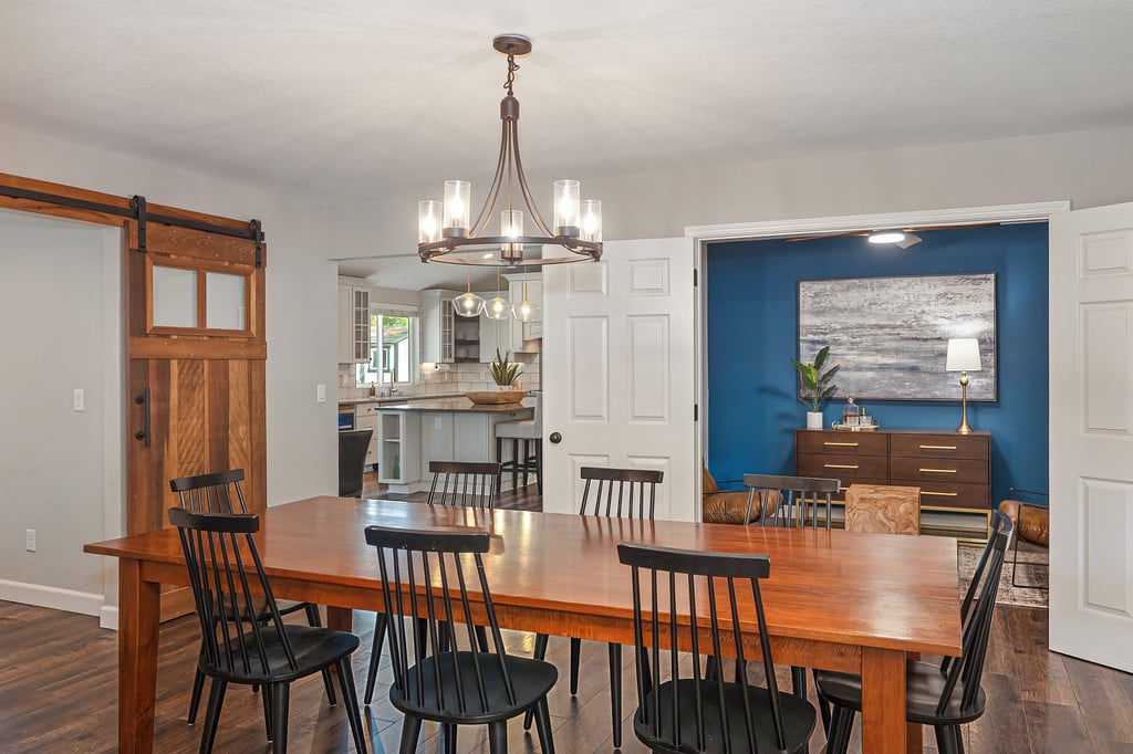 Dining room with a long wooden table, barn-style door, and blue-accented sitting room beyond.