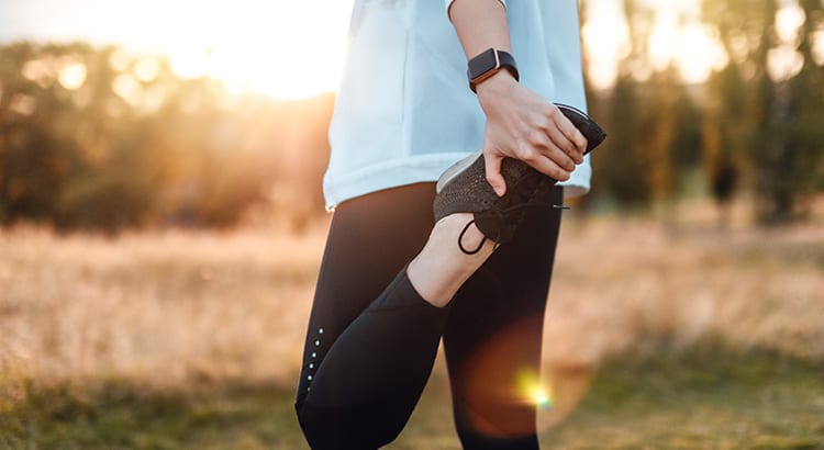 Woman with a pale blue three quarter sleeve top, black leggings, and Apple Watch stretching her left leg back against her thigh