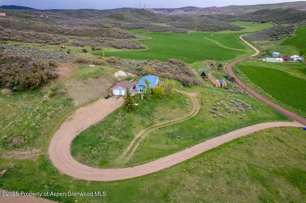 Aerial view of a rural home beside a winding dirt road, surrounded by green fields and rolling hills.