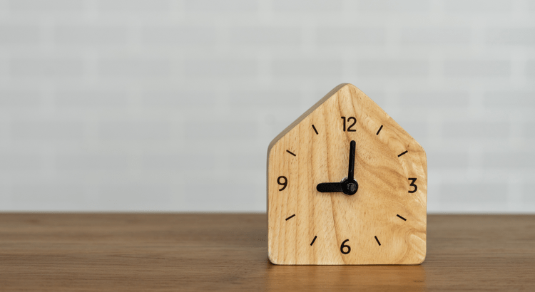 Wooden clock shaped like a house on a wooden table. Simple black hands show 10:10 against a light background, conveying a minimalist, serene tone.