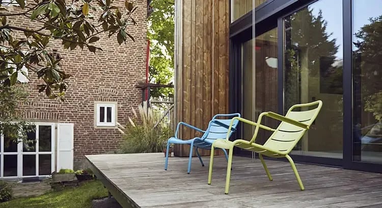 There are two chairs and a table on a wooden deck, with a view of a green backyard and part of a brick house. The scene suggests relaxation and enjoying nature.