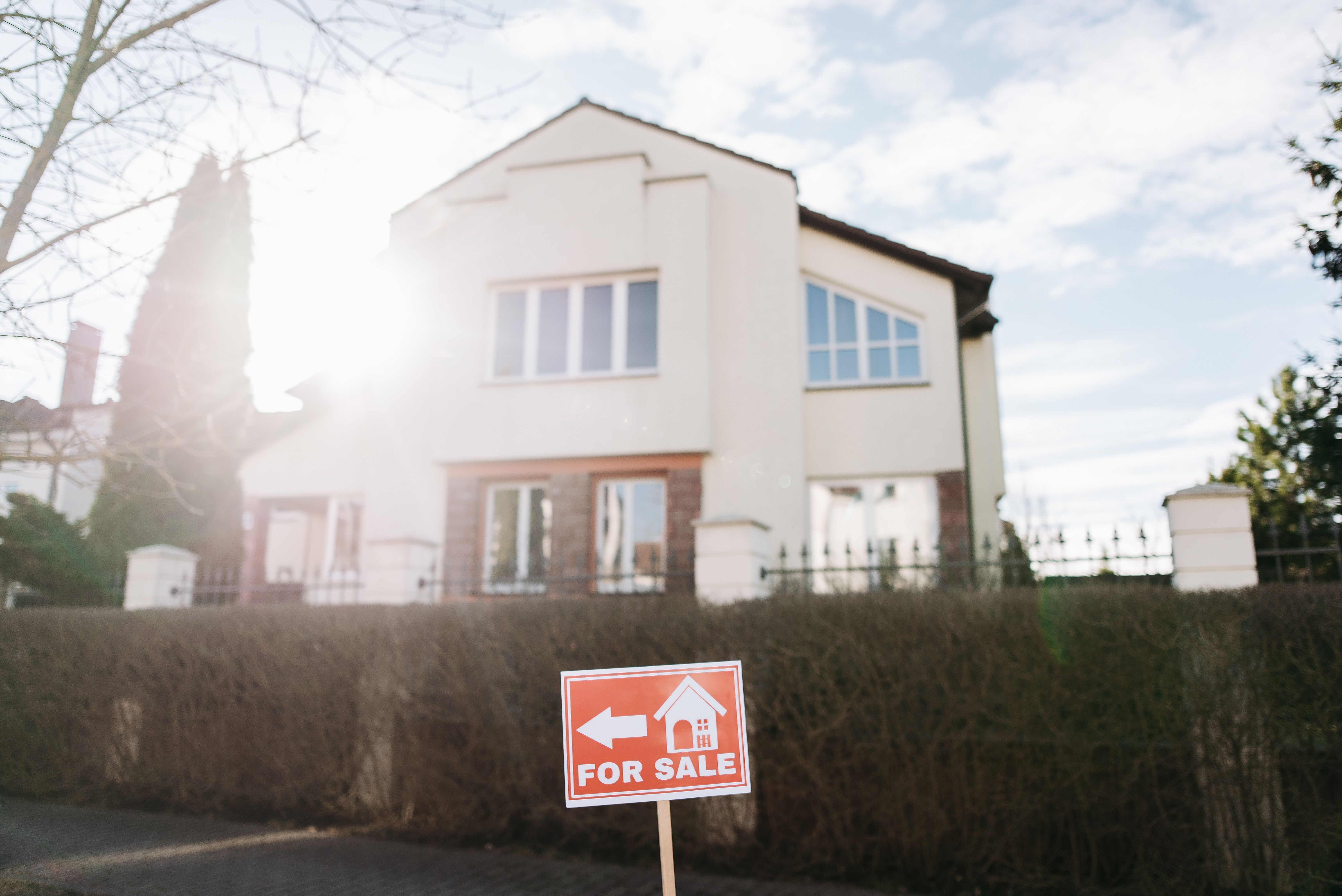 White two-story house behind a hedge, with a red-and-white for-sale sign in bright sunlight.