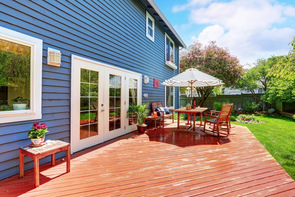 Backyard deck with outdoor dining set beneath a striped umbrella beside a blue house.