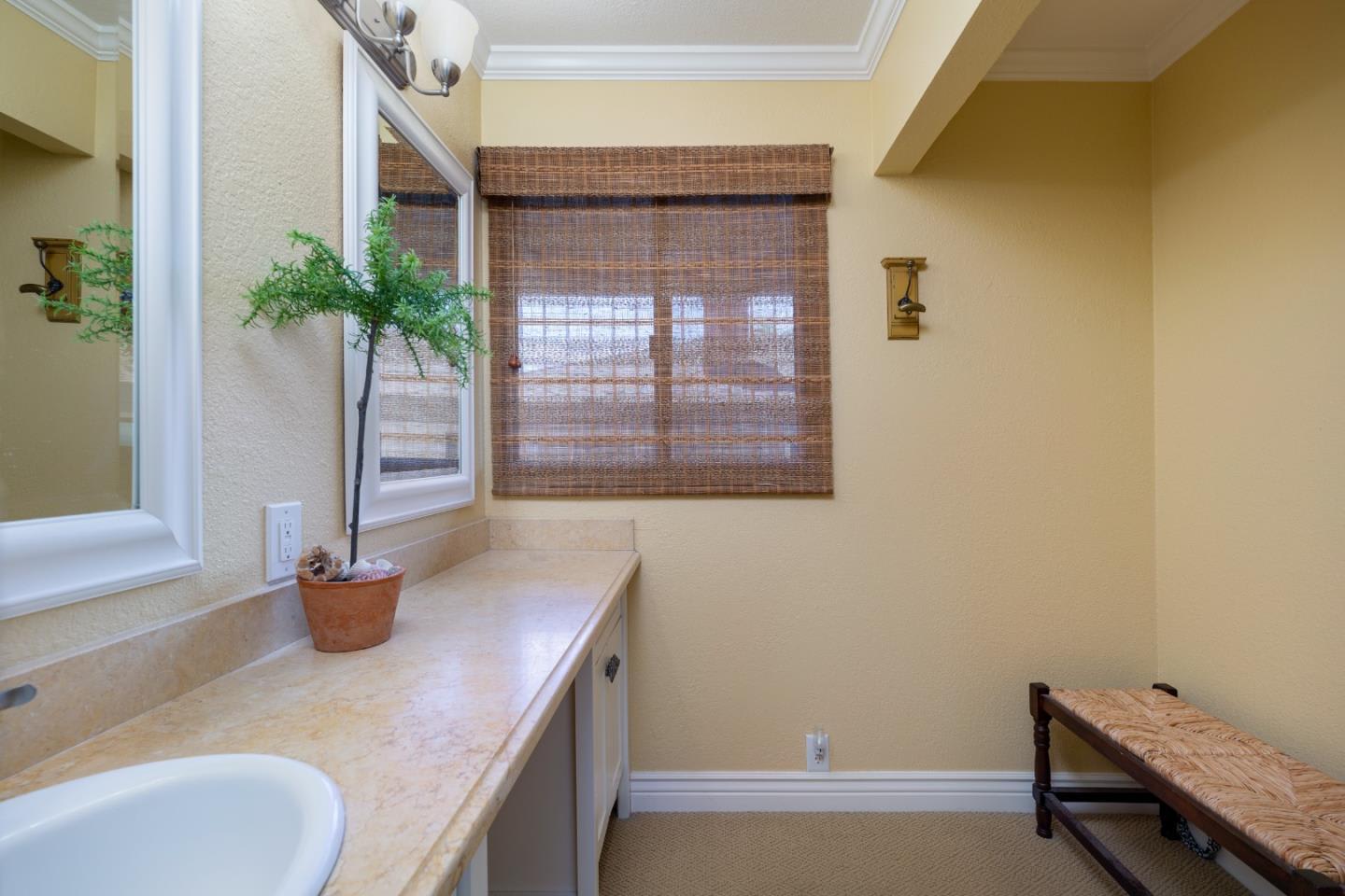 Bathroom vanity with a beige stone counter, large mirrors, woven shades, and an upholstered bench.