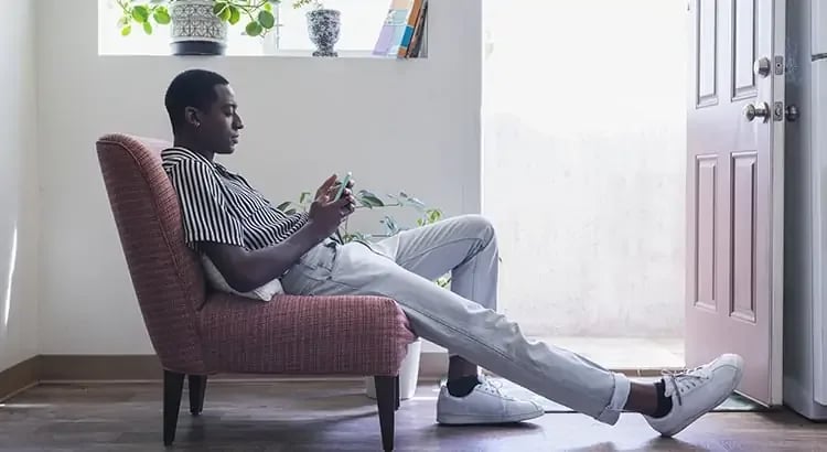 A man sitting in a chair near a door, holding his phone. The setting appears relaxed and comfortable.