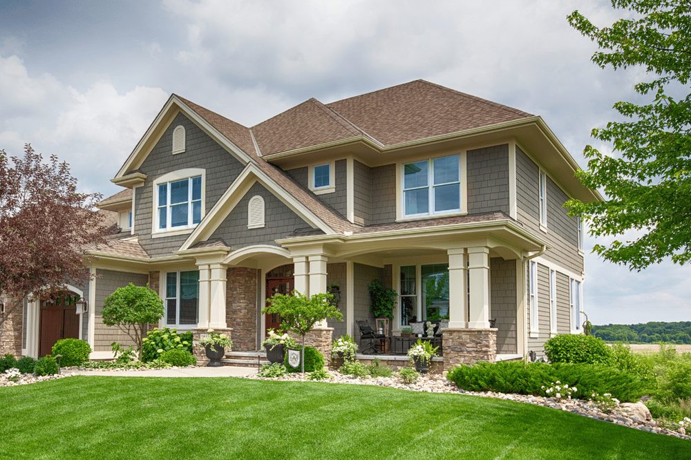 Two-story gray-shingled home with stone accents, a covered porch, and landscaped lawn.