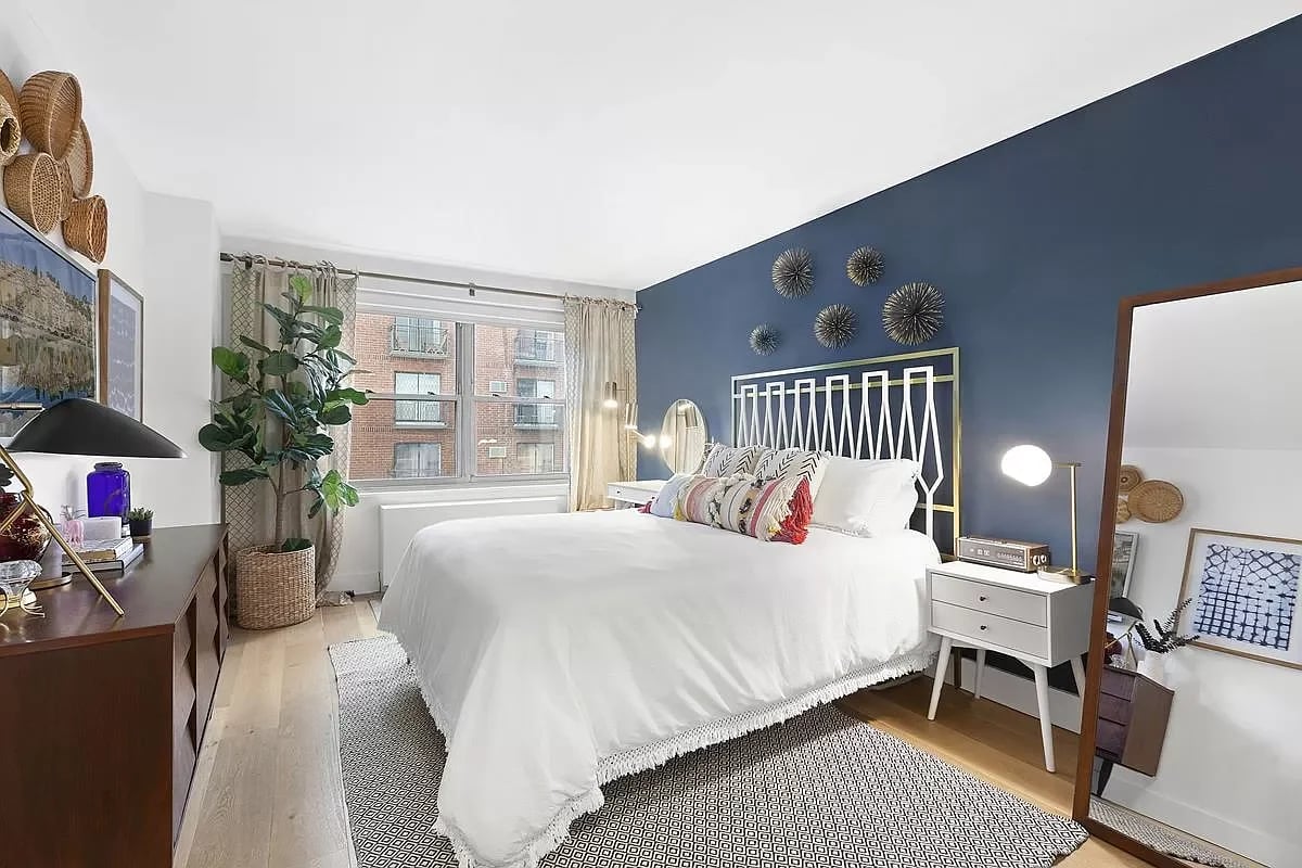 Bedroom with a white bed against a deep blue wall and a window overlooking brick buildings.