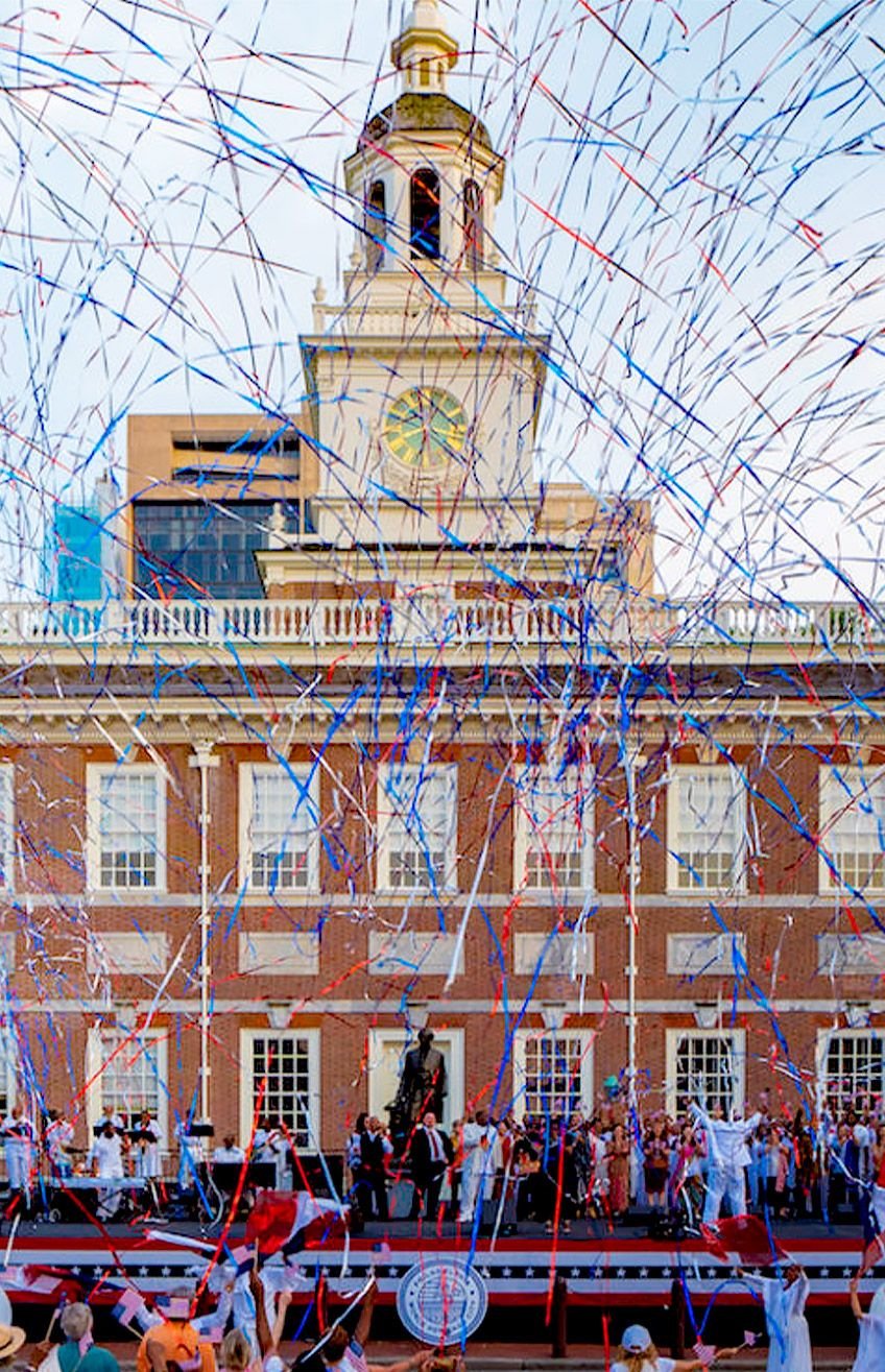 Independence Day parade crowd beneath red, white, and blue streamers in front of a clock-tower building.