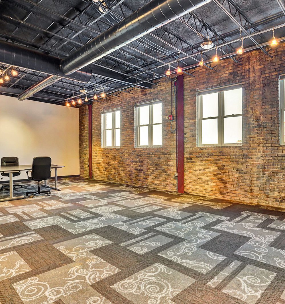 Office interior with exposed brick, ceiling ducts, patterned carpet, and a small meeting table.