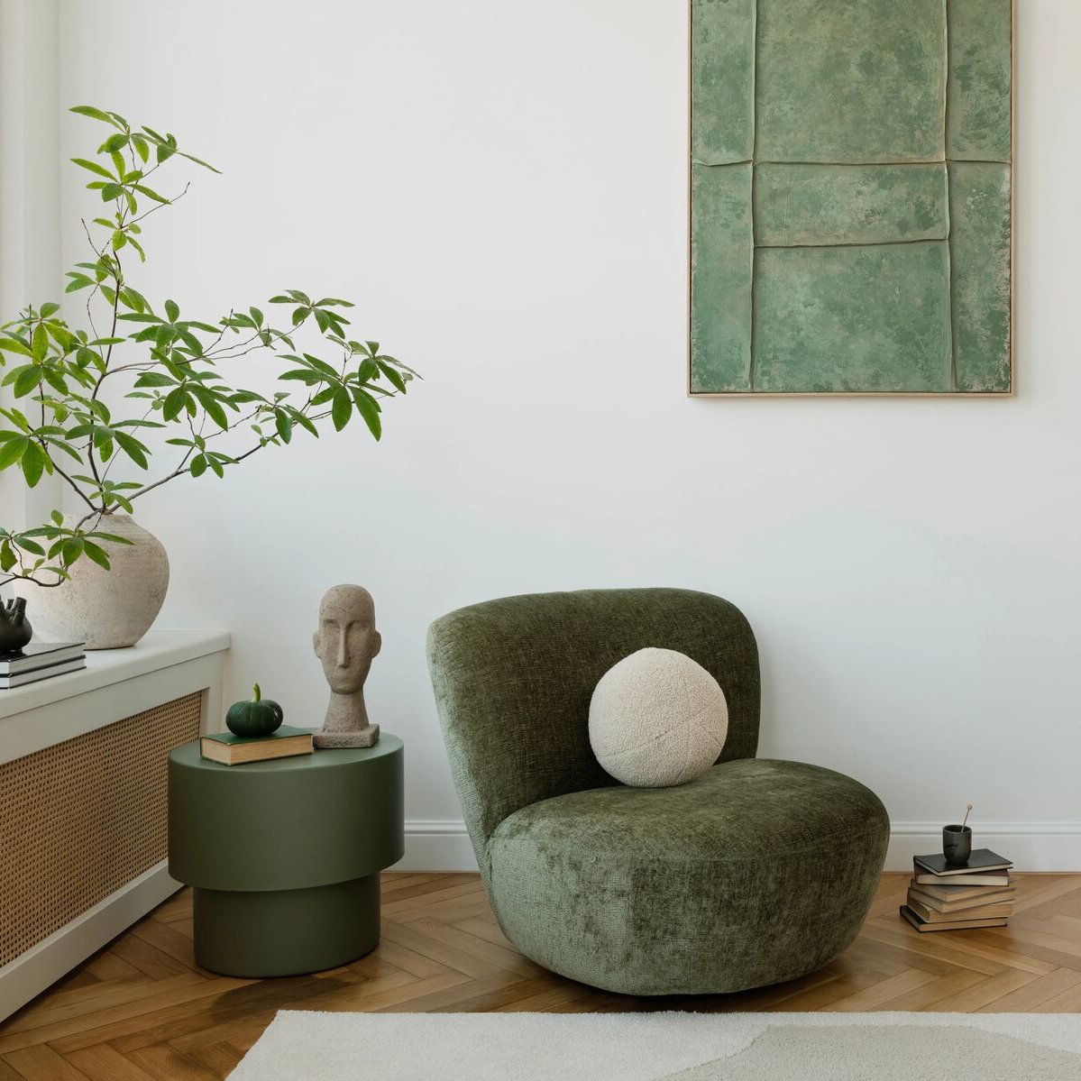 Living room with an olive-green chair, leafy plant, framed artwork, and herringbone wood floor.