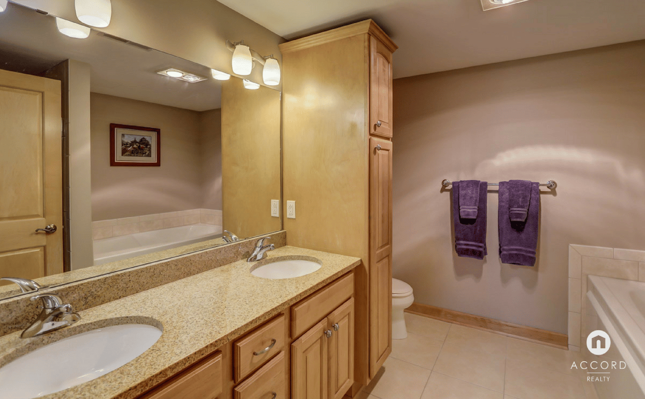 Condo bathroom with a double-sink vanity, speckled countertop, light wood cabinetry, and purple towels.
