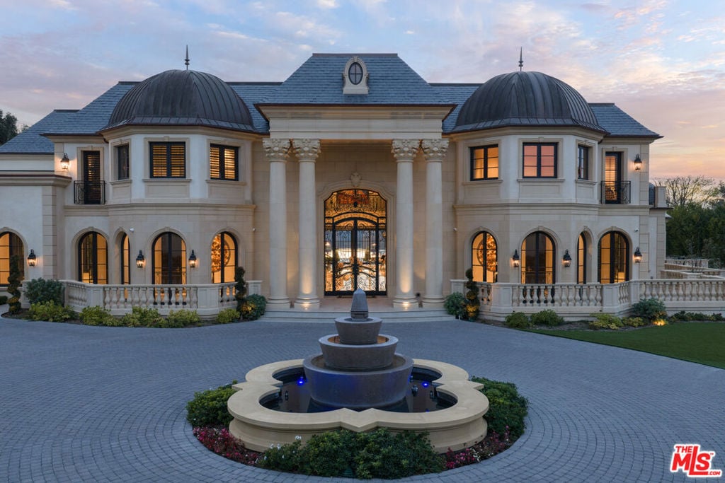 Mansion with twin domed roofs, columned entry, and tiered courtyard fountain at dusk.
