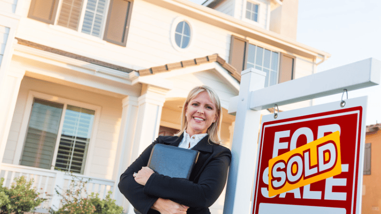 A real estate agent stands beside a SOLD sign in front of a large house.