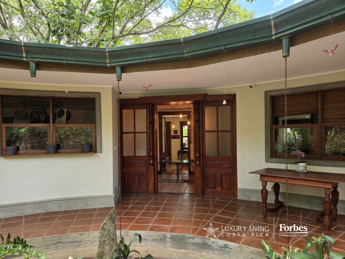 Covered tiled patio with wooden entry doors, a small table, and mature trees overhead.