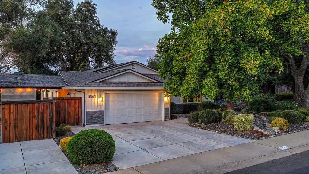 Single-story home with a two-car garage and broad driveway, framed by mature trees and landscaped shrubs.