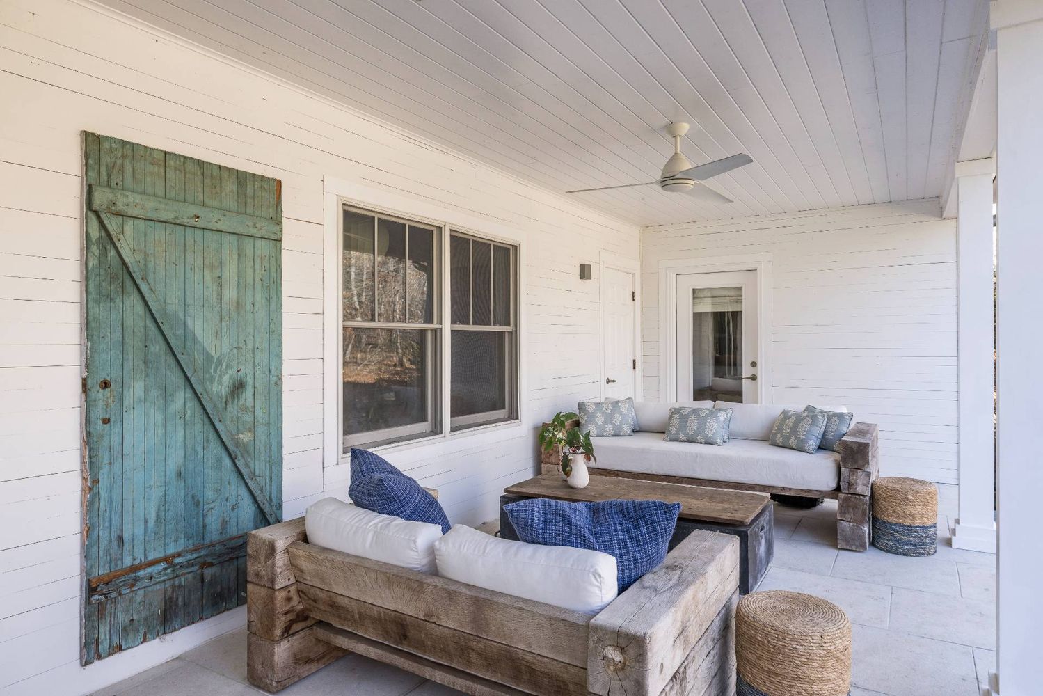 Covered porch with rustic sofas, blue pillows, a ceiling fan, and a weathered teal barn door.