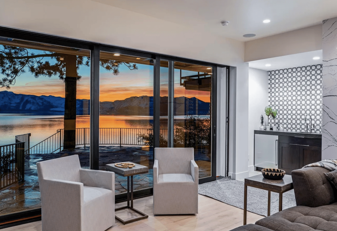 Modern living room overlooking Lake Tahoe and mountains at sunset through floor-to-ceiling glass doors.