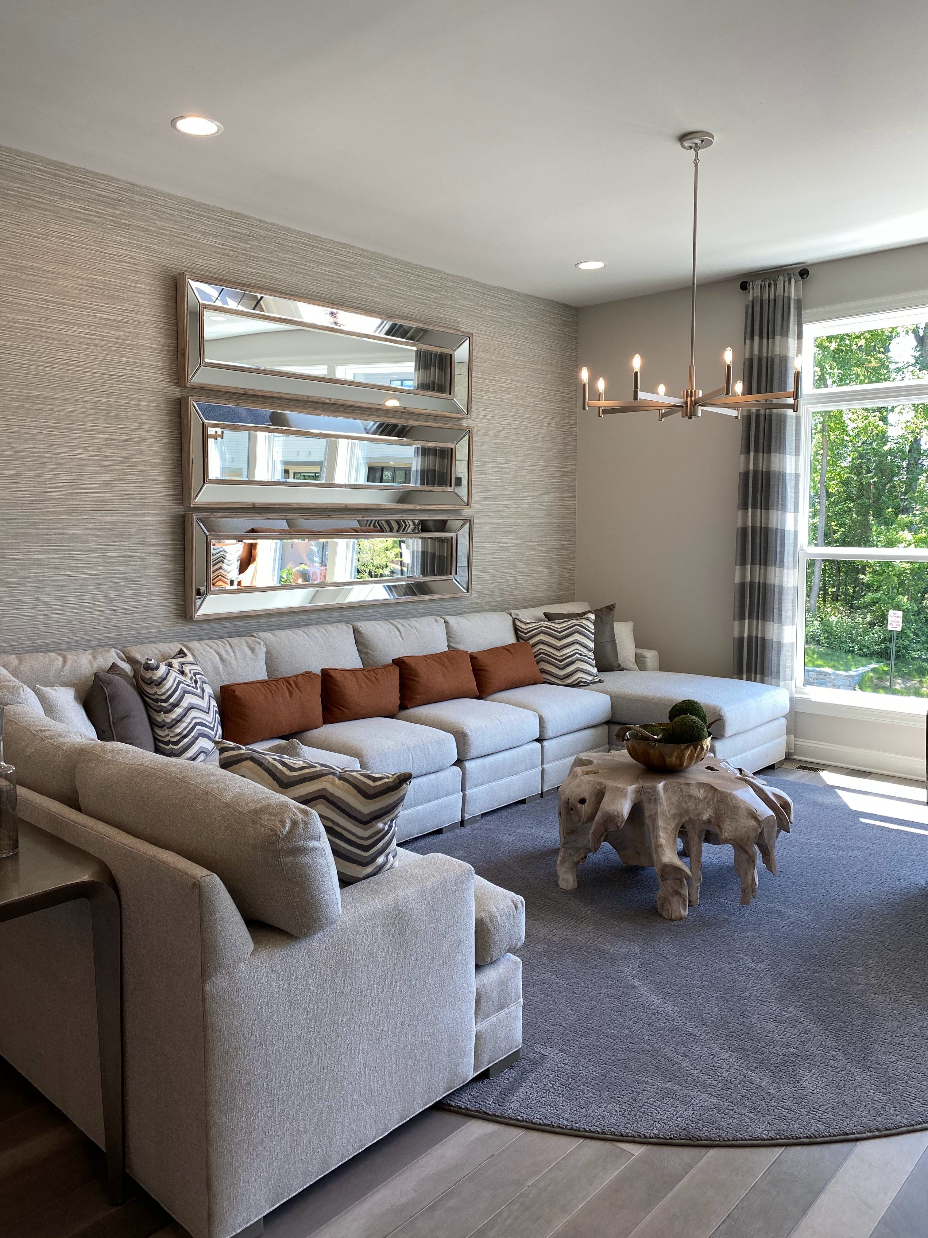 Staged living room with a gray sectional, rust pillows, three wall mirrors, and tall windows.