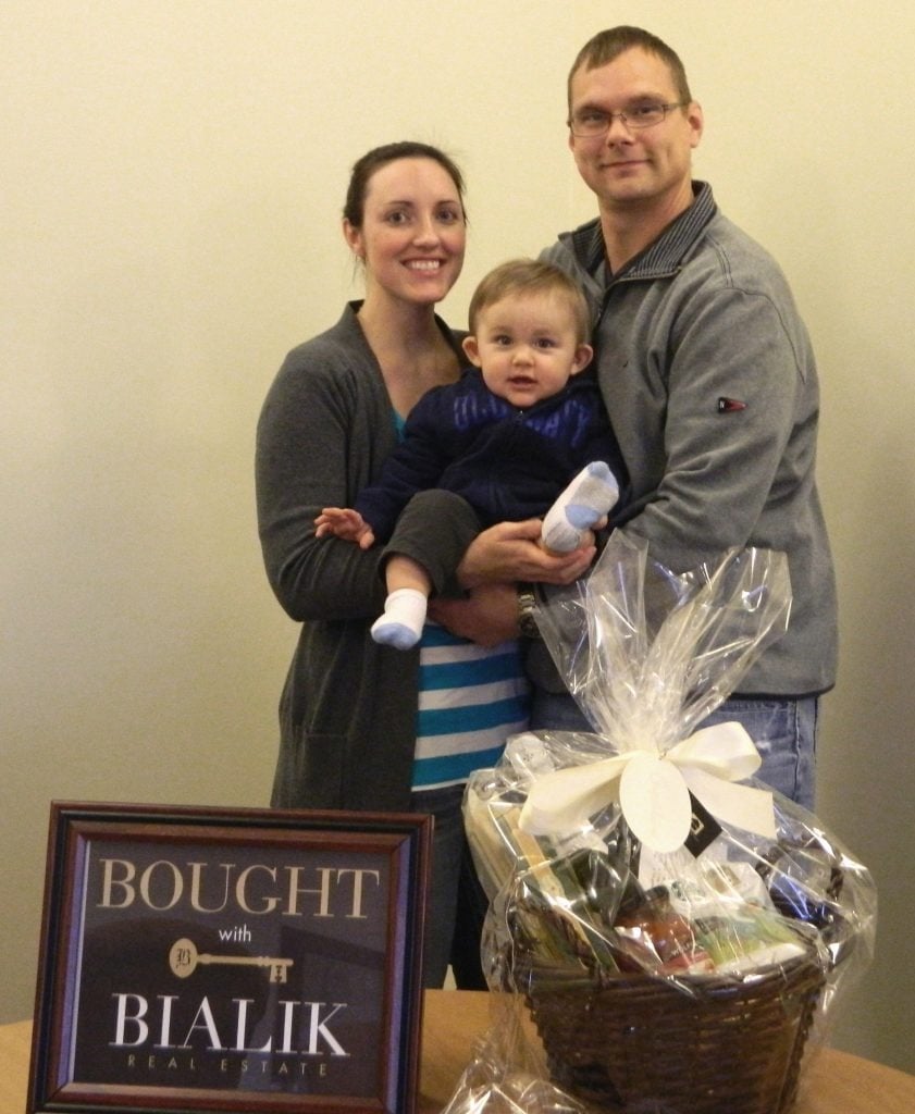 Jake and Kayla hold baby Grady beside a gift basket and a Bialik Real Estate sign.