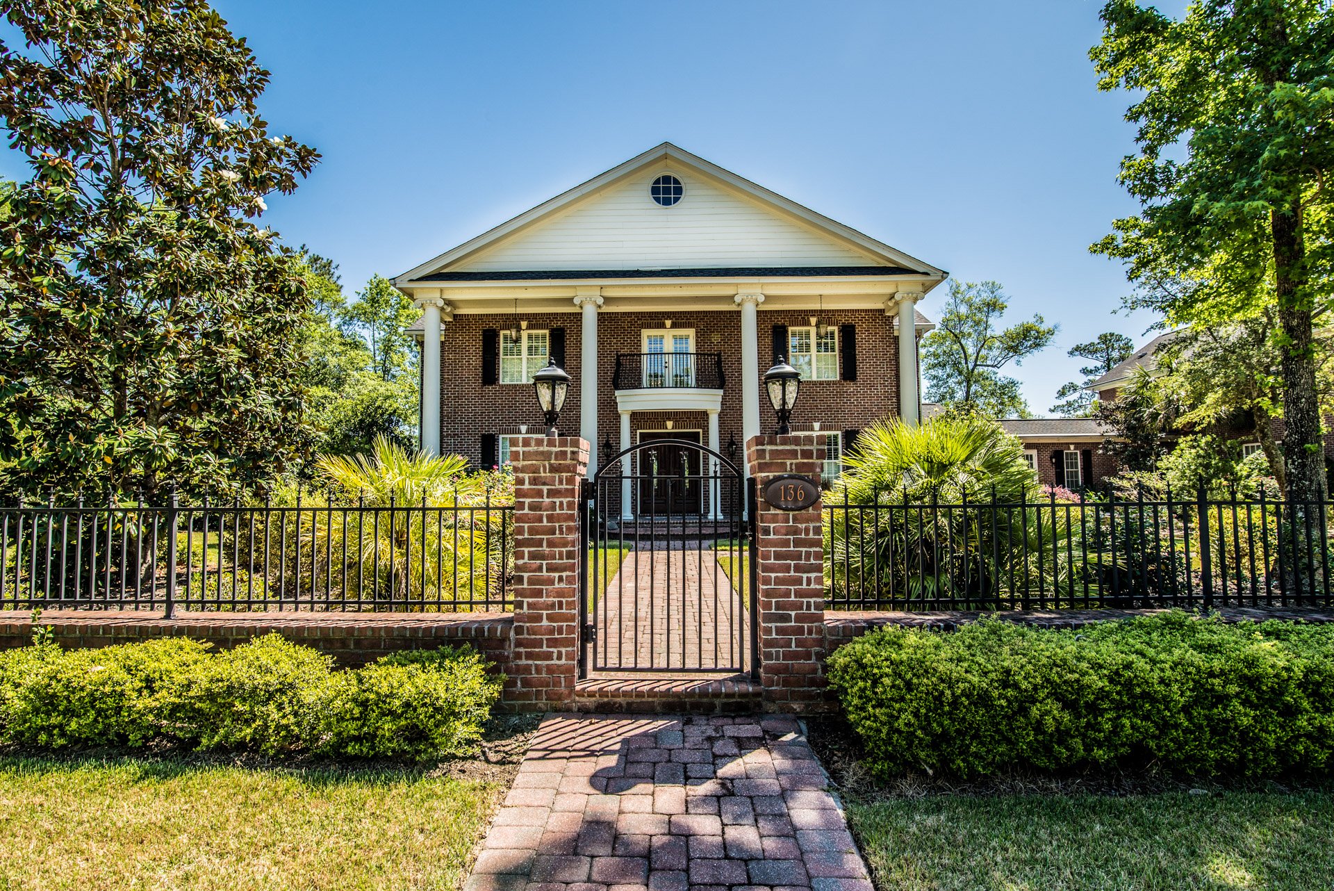 Two-story brick house with a columned porch behind a gated brick path and landscaped yard.