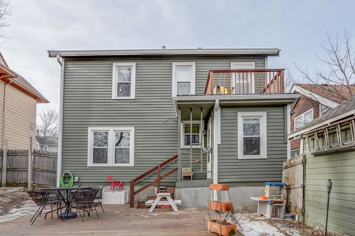 Rear exterior of a gray-sided home with a raised balcony, stairs, and patio seating.