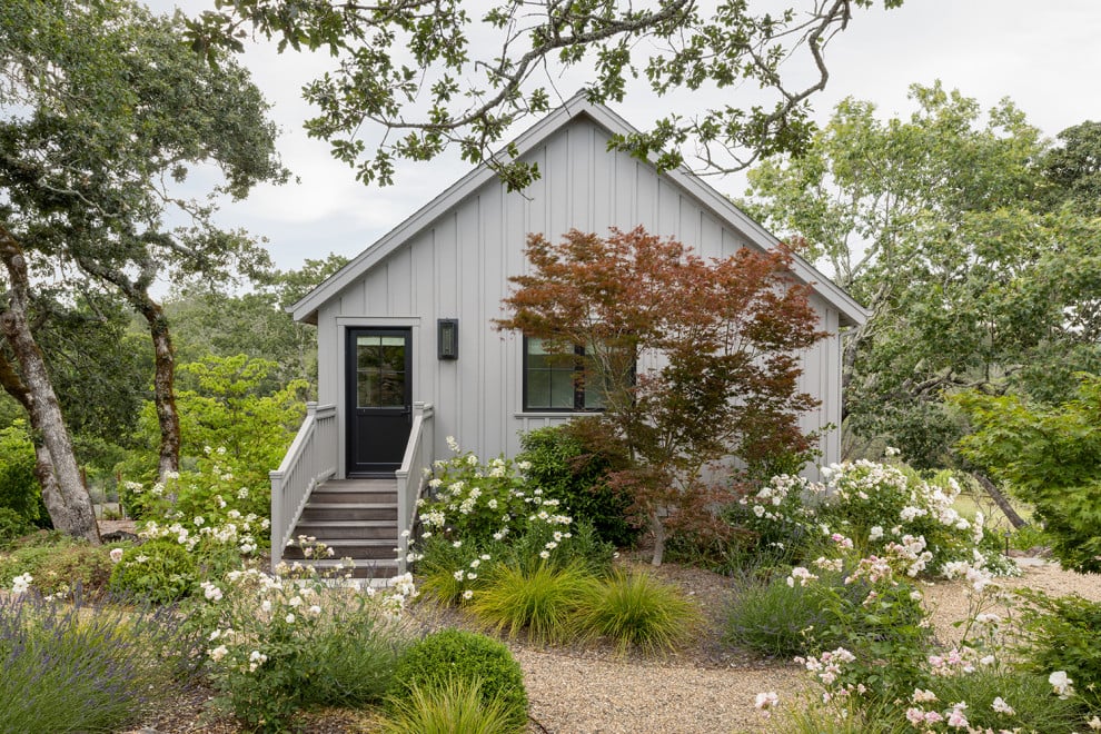 Light-gray cottage with a gabled roof, front steps, and a landscaped garden of flowering shrubs.