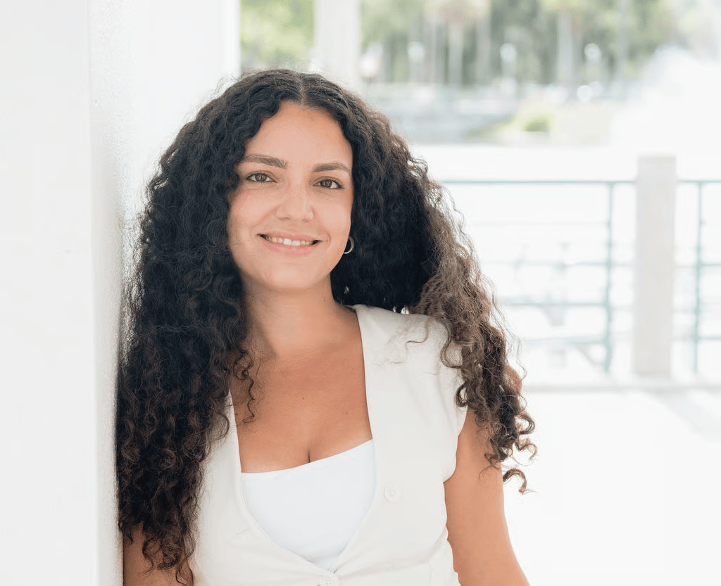 Smiling woman with long dark curls wearing a white sleeveless outfit.