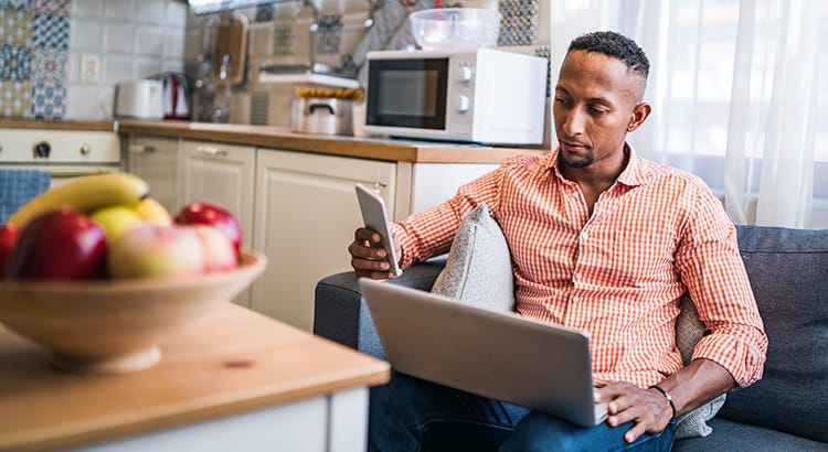 The man is sitting at the table, possibly engaged in remote work or personal tasks, with a notepad and pen in hand. A bowl of fruit is on the table, suggesting a homey atmosphere. This image conveys productivity, work-from-home, or a casual working lifestyle.