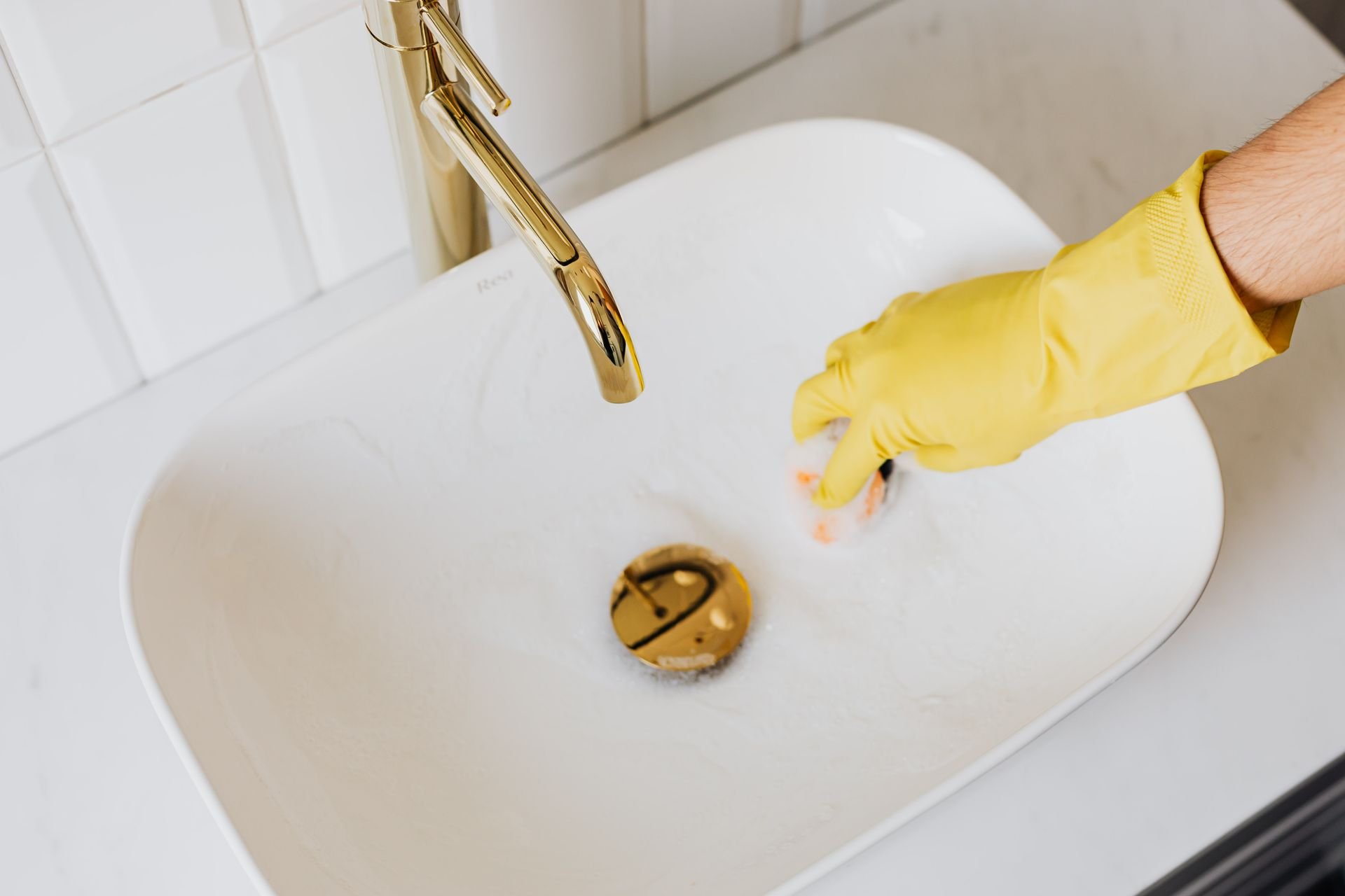 A yellow-gloved hand scrubs a white bathroom sink with a gold faucet.