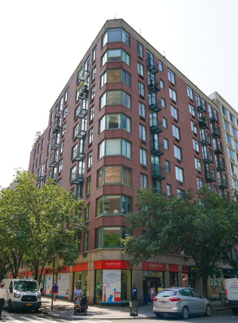 Red-brick corner apartment building with projecting windows, fire escapes, and ground-floor storefronts.