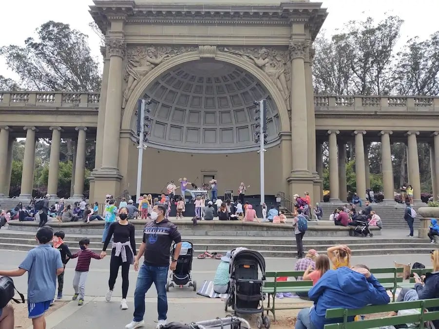 People picnicking on the grass while listening to the band, with the Bandshell in the background.