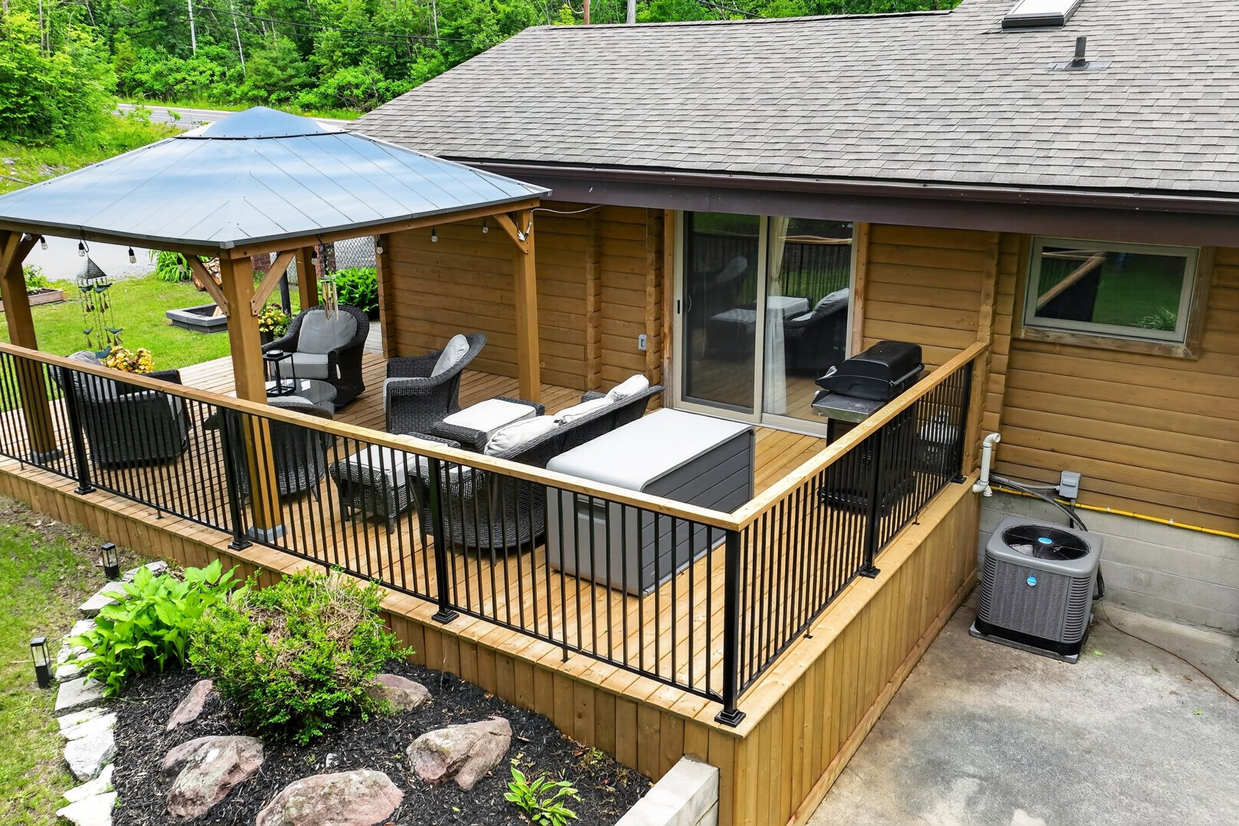 Furnished wooden deck with a covered gazebo, outdoor seating, and a barbecue beside a log-sided home.