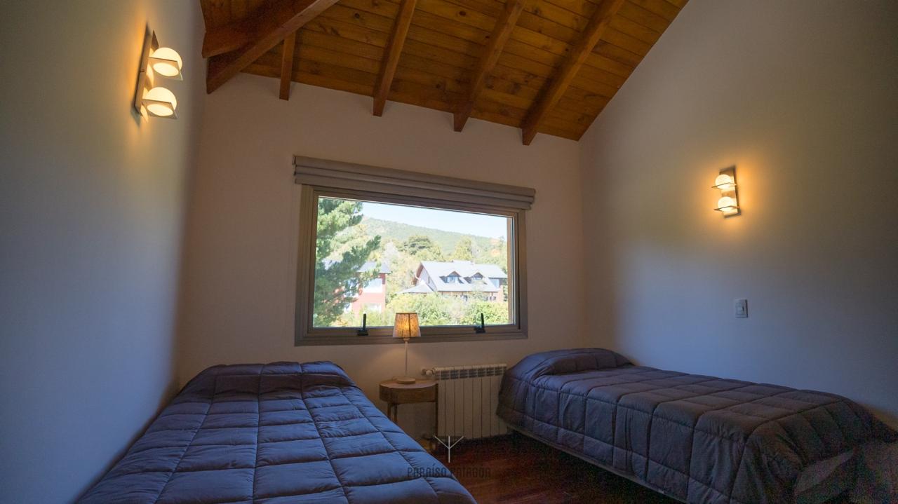Bedroom with two single beds beneath a vaulted wood ceiling and a window overlooking wooded hills.
