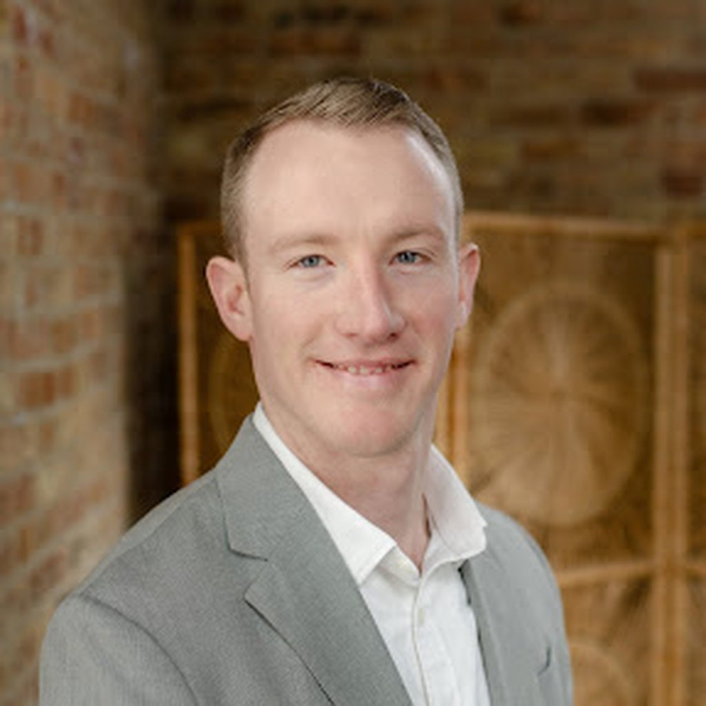 Man in a light gray suit and white shirt, smiling against a brick and wood background.